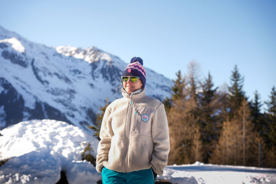 Woman wearing the Nanook23 stone recycled sherpa fleece with a smooth chest panel, turquoise zip pulls and the embroidered Mountains and Rivers badge on the left chest, with snowy mountains and trees behind.