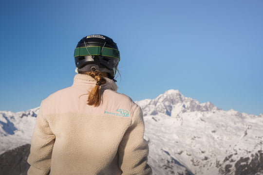 Back of the Nanook23 stone recycled sherpa fleece, with the Nanook23 name and geometric polar bear embroidered in turquoise on the right shoulder, worn with a ski helmet while looking out at a snow-covered mountain peak.