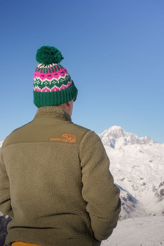 Back of the Nanook23 khaki recycled sherpa fleece, with the Nanook23 name and geometric polar bear embroidered in orange on the shoulder, worn while looking out at a snow-covered mountain peak.