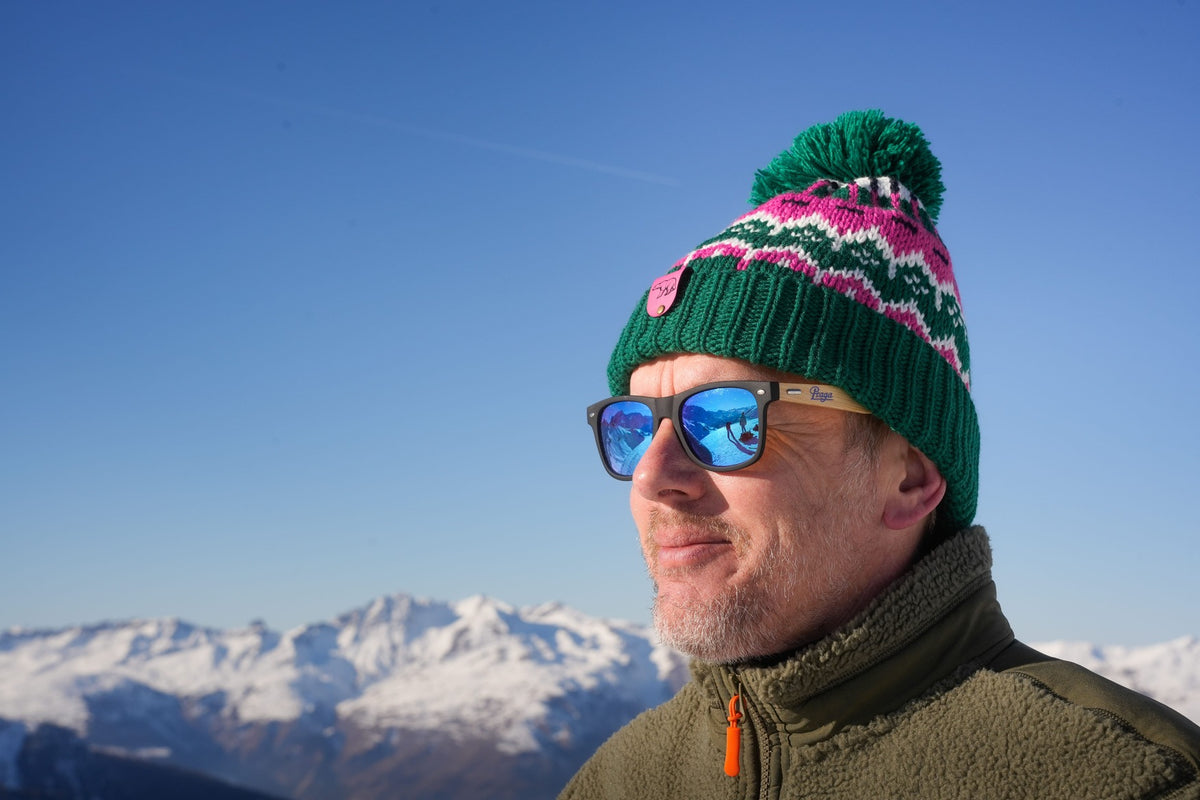 Man wearing the Nanook23 Alpine Trails recycled bobble hat in green, pink and white with a green pom-pom, alongside the khaki sherpa fleece, in front of a range of snow-capped mountains.