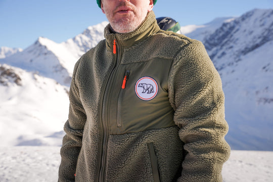 Person wearing the Nanook23 khaki recycled sherpa fleece with a smooth chest panel, orange zip pulls and an embroidered geometric polar bear patch on the chest, with snowy mountains and trees behind.
